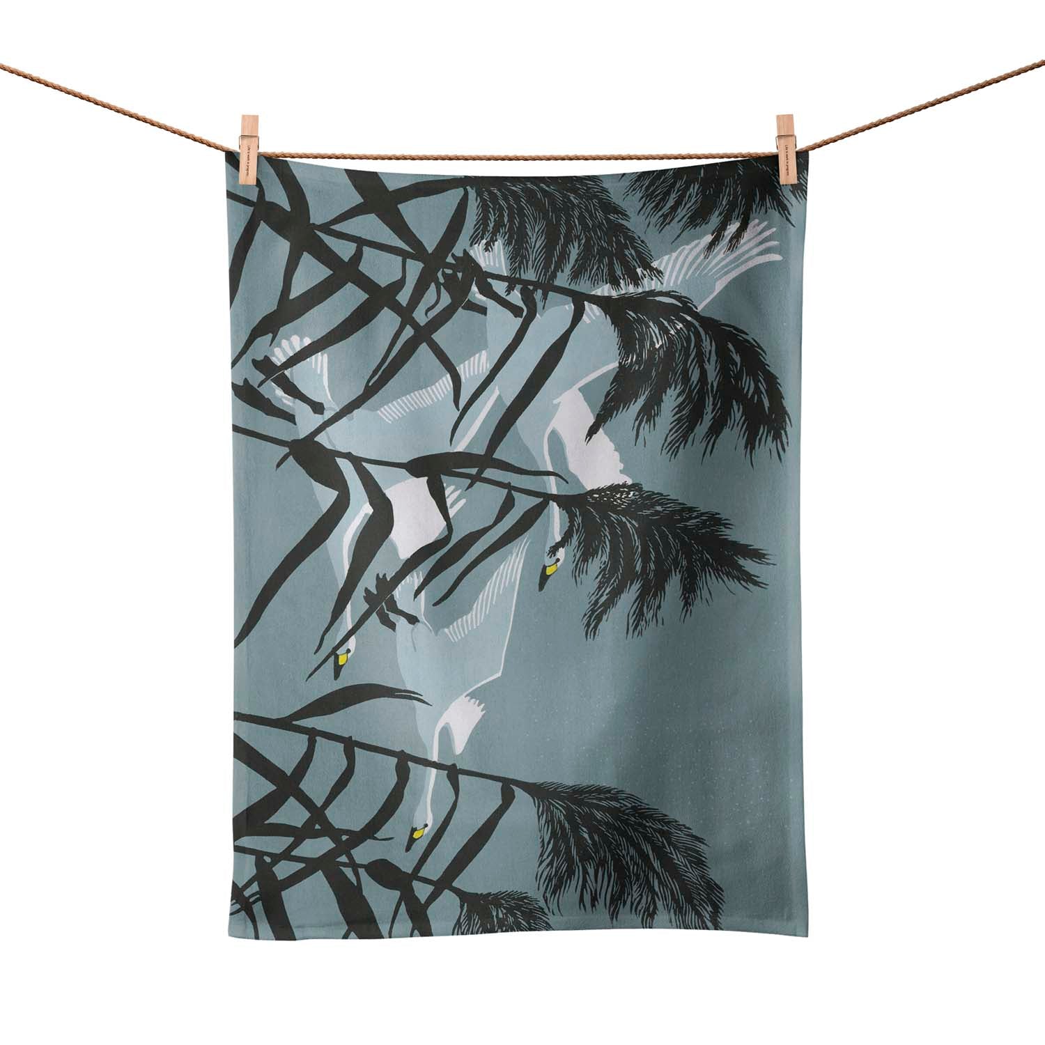 A grey/blue tea towel pegged on a washing line. Design is three swans   flying behind tall grasses.